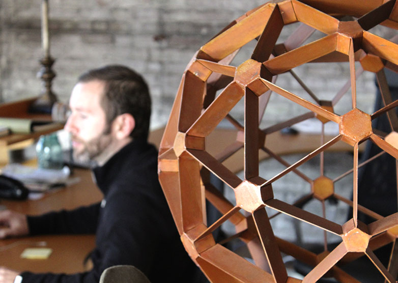 A man works at a computer in soft focus, behind a spherical abstract sculpture.