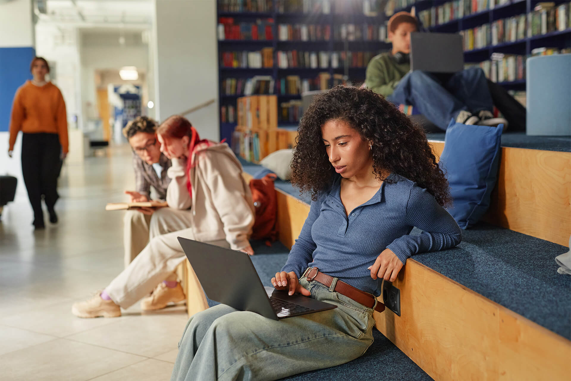 Young adult woman sitting on steps using laptop, surrounded by diverse group of young adults and teenagers studying with digital devices in modern library