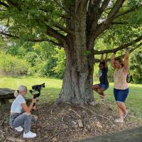 Photo of a young woman taking video of a girl and a woman hanging from a tree brnach