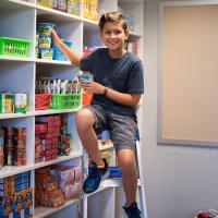 photo of a boy siting in from of a shelve containing food supplies