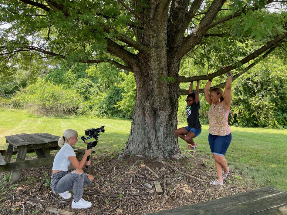 Photo of a young woman taking video of a girl and a woman hanging from a tree brnach